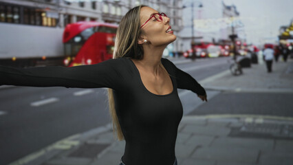 Fototapeta premium Young woman smiling with arms outstretched on a busy city street beside red double decker bus, looking upward at the sky; joy exploration.