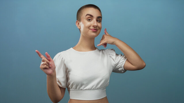 Woman pointing left with index finger and making a thumb to ear phone gesture, showing exposed midriff and puffy sleeve white crop top in a studio; playful mood.