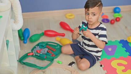 Fototapeta premium Little boy playing with green toolkit and toy car, holding pieces on foam alphabet mat inside building; concentration.