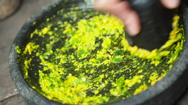 Close-up of hand grinding green chili and yellow spice paste in a traditional stone mortar with pestle, preparing fresh sambal or curry base