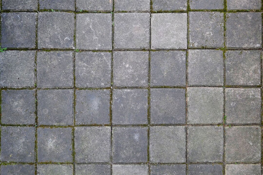 Weathered square concrete paving block texture with natural gray stone tones, visible grout lines, and subtle moss between joints. Ideal for outdoor pavements, walkways, landscape design, architectura