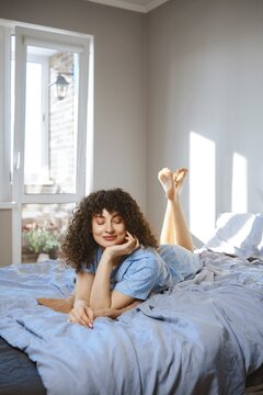 A young woman is lying on her stomach on a bed covered with blue sheet. She rests her chin on her hand and smiles while looking thoughtful. Sunlight streams through a nearby window
