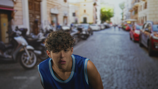 Teenage boy wearing los angeles basketball jersey grimaces with eyes closed while leaning forward on a cobbled street; awkward discomfort.