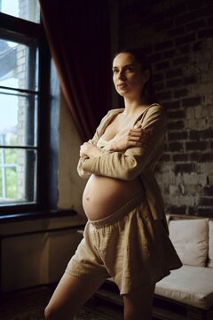 A pregnant woman stands at home, wearing comfortable attire. The cozy room features large windows allowing natural light, showcasing her glowing anticipation of motherhood