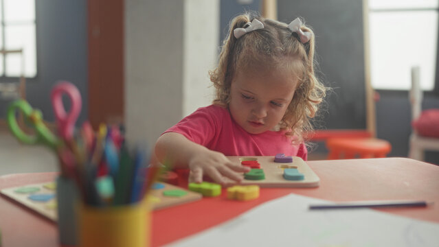 Girl places a yellow puzzle piece with her right hand on a wooden board at a classroom table, focused on shapes and colors; concentration learning curiosity.