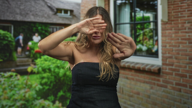 Woman shields eyes with hands and holds palms forward in front of brick building window and garden; shyness privacy solitude.