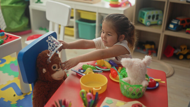 Hispanic girl reaches with both hands to place plastic tiara on a stuffed monkey at a playroom building; imaginative joy.