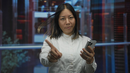 Fototapeta premium Woman tapping smartphone with hands in hotel atrium by glass railing while checking messages and pointing finger at screen; calm concentration.