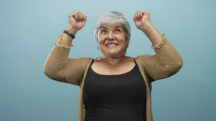 Woman raising arms and smiling with exposed shoulders and hoop earrings against a blue studio backdrop, wearing a cardigan and tanktop  joy celebration. © Krakenimages.com