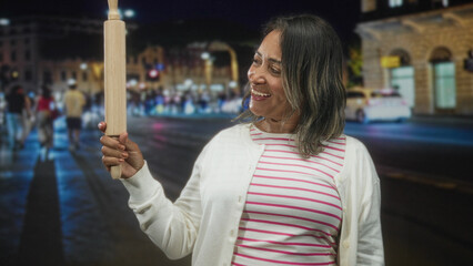 Woman smiling holds rolling pin upright with right hand wearing striped shirt and cardigan on a busy city street at night  joyful confidence. © Krakenimages.com