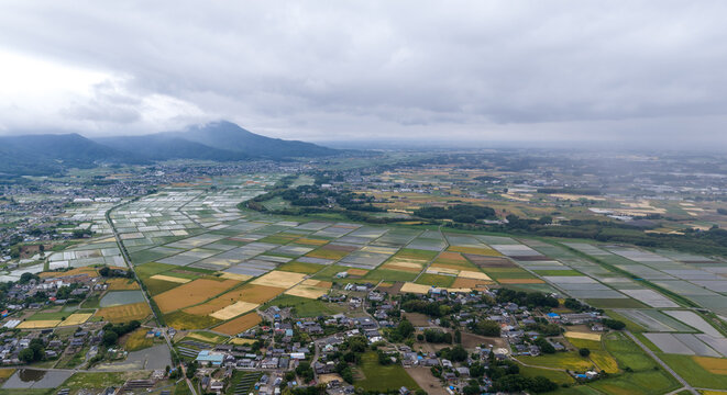 Aerial view of the Iwase Rice fields featuring a vast patchwork of agricultural paddies nestled against a backdrop of rolling mountains under a cloudy sky, Iwase, Ibaraki, Japan.