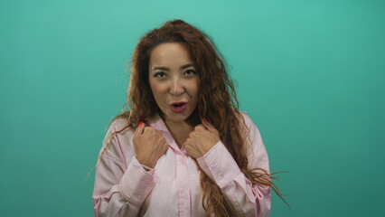 Fototapeta premium Woman young caucasian clenching fists to chest, hands visible with red nails and open mouth, long curly hair, posed against a teal studio wall; excitement.