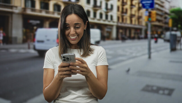 Woman holding smartphone and tapping screen, smiling with a small fist gesture on a city street in front of buildings and vans; joy connection.