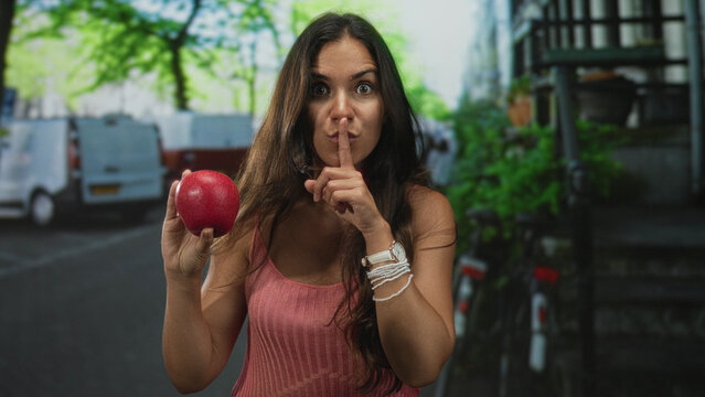 Young hispanic woman holding a red apple, finger to lips for silence on an urban street with parked vans; playful secrecy.