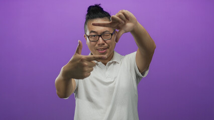 Fototapeta premium Man framing with hands and winking eye in purple studio set wearing white polo and glasses with top knot; playful creativity.