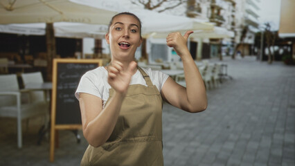 Fototapeta premium Woman in apron gestures with hands and points thumbs at a chalkboard menu outside a street cafe patio; playful communication.