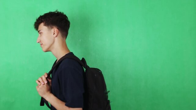 Young male student with black backpack stands in profile against a green backdrop, showcasing a casual pose with hands clasped in front of him, emphasizing a relaxed demeanor