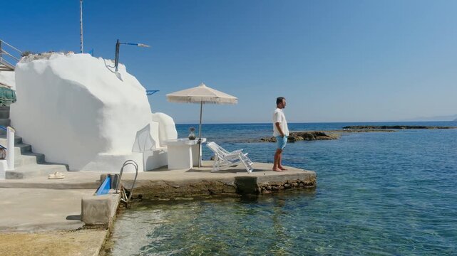 Breathtaking coastal vistas of Mandrakia Milos island showcase crystal-clear waters, inviting charm, and tranquil moments under the bright sun. A man relaxing in front of a local fisherman hut