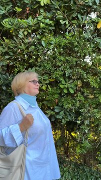 Senior woman walking with a shoulder bag in a quiet green urban area. Older woman in casual clothing walking outside during the day.