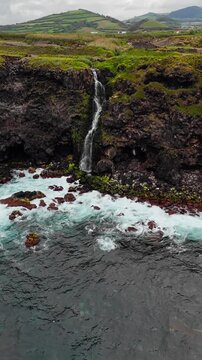 Aerial view of waterfall flowing down volcanic cliff into Atlantic Ocean on Azores coast, dark rocks and ocean waves under cloudy sky