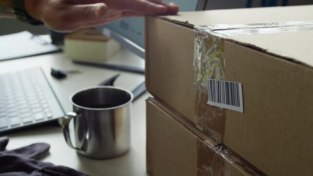 Cropped shot of warehouse worker scanning barcode on cardboard box using handheld laser scanner and reaching for metal mug. Footage is perfect for e-commerce, logistics, and shipping commercials