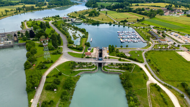 Aerial view of a river lock system and a small marina with docked boats, surrounded by lush green fields and trees in Auvergne-Rhone-Alpes, France.