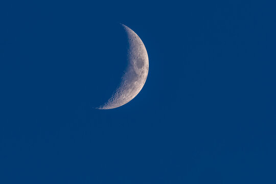 Close up of a waxing crescent moon with detailed craters against a deep blue sky background.
