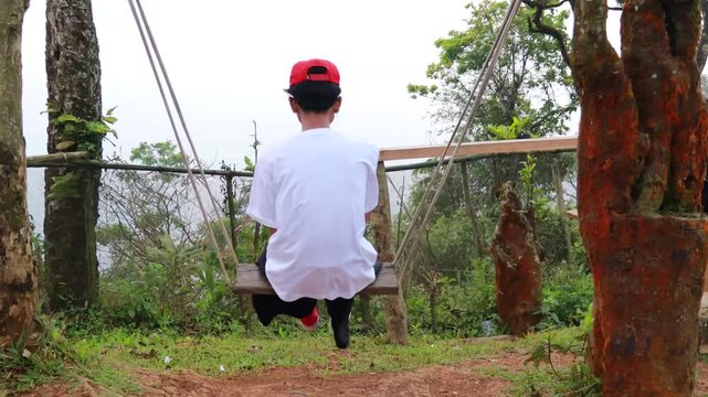 Young man in white shirt and red cap sitting on a wooden swing.