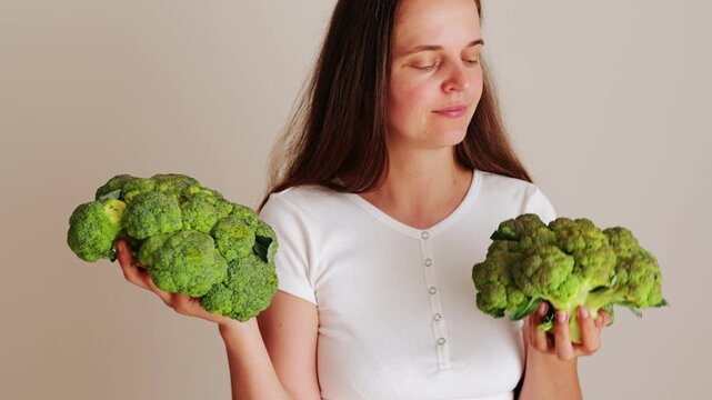 A woman stands in a kitchen holding two heads of broccoli. She looks carefully at both vegetables, deciding which is better. Natural light comes through a window