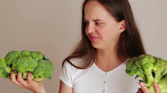 A woman stands holding two heads of broccoli, looking thoughtfully at them. The background is plain, and she wears a white shirt. Natural light fills the space