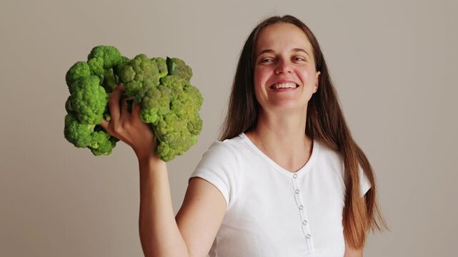 A woman is laughing and showing off her strength by holding a big bunch of broccoli. She is enjoying a lighthearted moment in a simple indoor setting