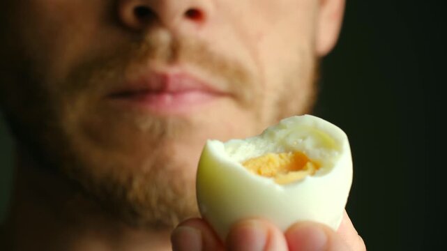 Close up of bearded man chewing a bitten hard boiled egg. Focus on half eaten egg with visible bite mark in his mouth. Bearded man eating healthy hard boiled egg 