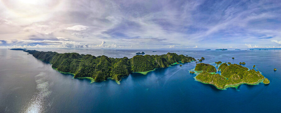 Aerial view of Raja Ampat Regency featuring lush green karst islands surrounded by turquoise waters and coral reefs under a bright sky in Southwest Papua, Indonesia.