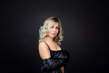 Fototapeta premium A blonde woman in a black leather jacket and blue jeans is standing in a dark studio. She is wearing a black bra and has her beautiful breasts exposed.