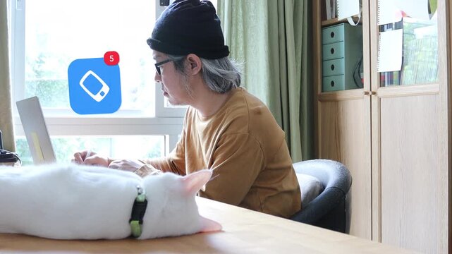 Side view of a mature Asian man using a laptop at home office next to his cat, displaying a 3D tracked smartphone mobile device network notification pop up and red counter.