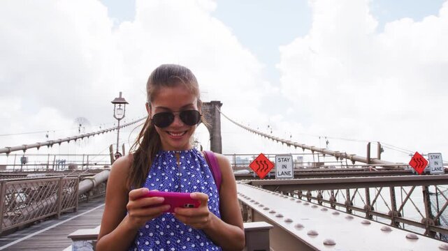 Happy Asian woman using phone texting walking on Brooklyn bridge, New York city for social media or blogging. Tourist doing summer travel in Manhattan, USA.