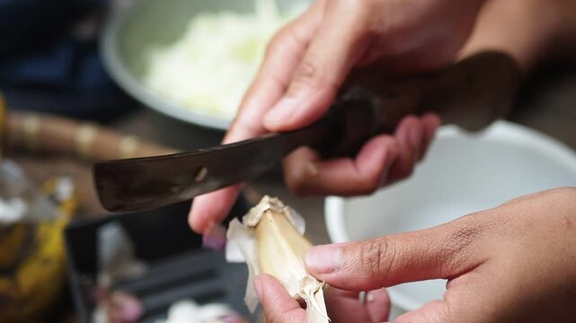 Close-up of hands peeling garlic cloves with a knife during food preparation in a rustic kitchen setting.