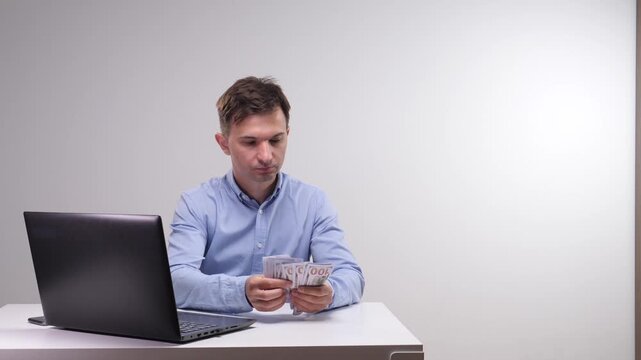 Man in a blue shirt sitting at a white desk. Carefully counting a stack of one hundred dollar bills next to an open laptop computer. Representing concepts of finance. Income. Payment