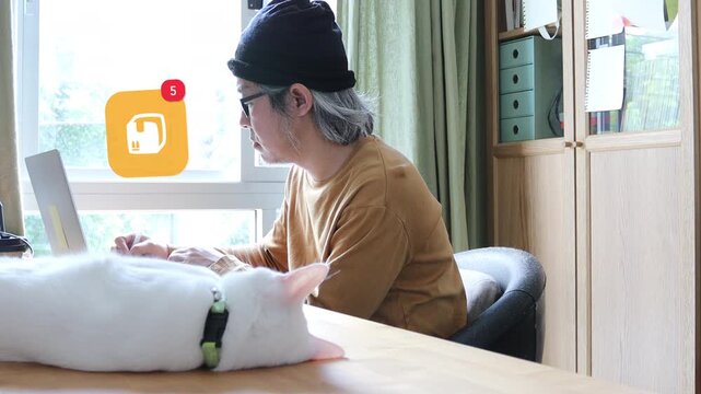 Side view of a mature Asian man using a laptop at home office next to his cat, displaying a 3D tracked parcel box logistics shipping notification pop up and red counter.