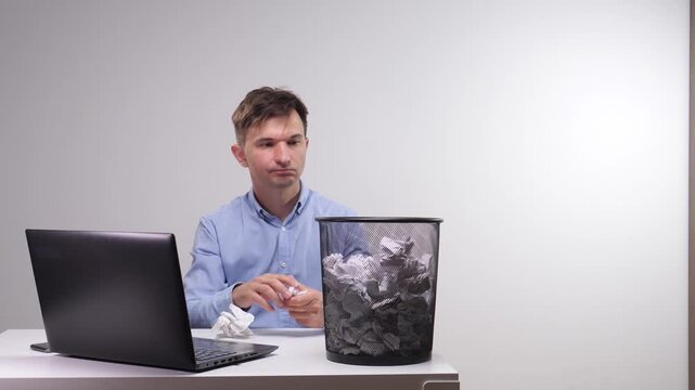 Man experiences creative block, frustration, and stress, crumpling paper and discarding it into a wastebasket after failing to complete work at a desk with a laptop