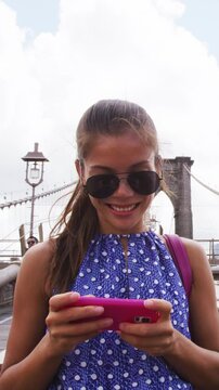 Happy Asian woman using phone texting walking on Brooklyn bridge, New York city for social media or blogging. Tourist doing summer travel in Manhattan, USA. Vertical Video.