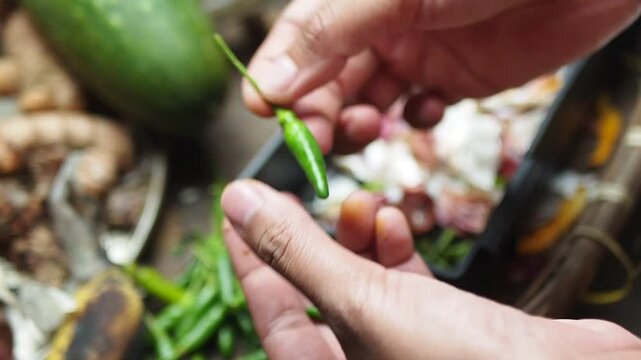 Close-up of hands plucking stems from fresh green chilies, preparing ingredients for cooking, with organic waste being collected in a compost bin.