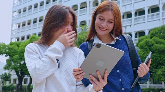 Happy female student and classmate share digital tablet on university campus for education, smiling young girl friend learning outdoor together near school building during study break
