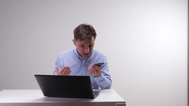 Young man reacting with distress and anger while looking at his smartphone, working at a laptop on a white desk
