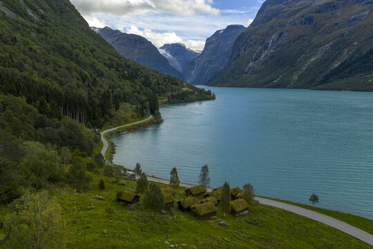 Aerial view of the historic Breng seter farm with traditional grass-roofed cabins on the lush green shores of the turquoise Lake Lovatnet, Stryn Municipality, Vestland, Norway.