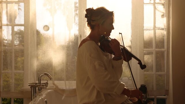 young woman playing violin in bathroom at sunset