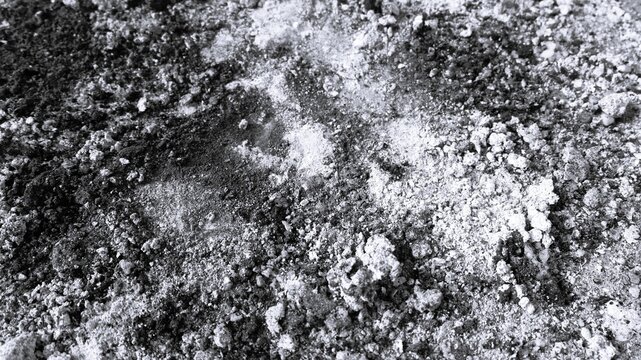 Black and white close-up of coarse dusty rubble with mixed powder patches, rough granules, and abstract construction texture.