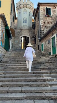 Senior woman walking up old stone steps toward a clock tower in a historic European town. Mature tourist sightseeing in a Mediterranean old town.
