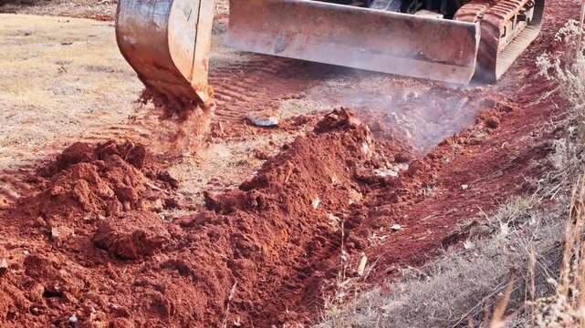 Bulldozer clears red soil in field, preparing ground for future construction, dust rises from work site.