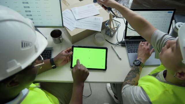 High angle view of African American male worker in hard hat using green screen tablet with young female colleague typing on laptop at warehouse desk. Ideal for logistics software promo